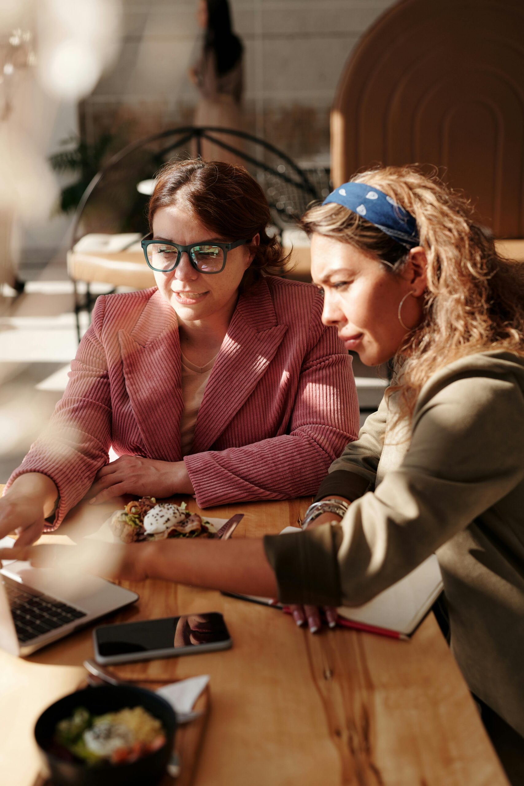 Two women engaged in teamwork at a café table, using a laptop in the sunlight.